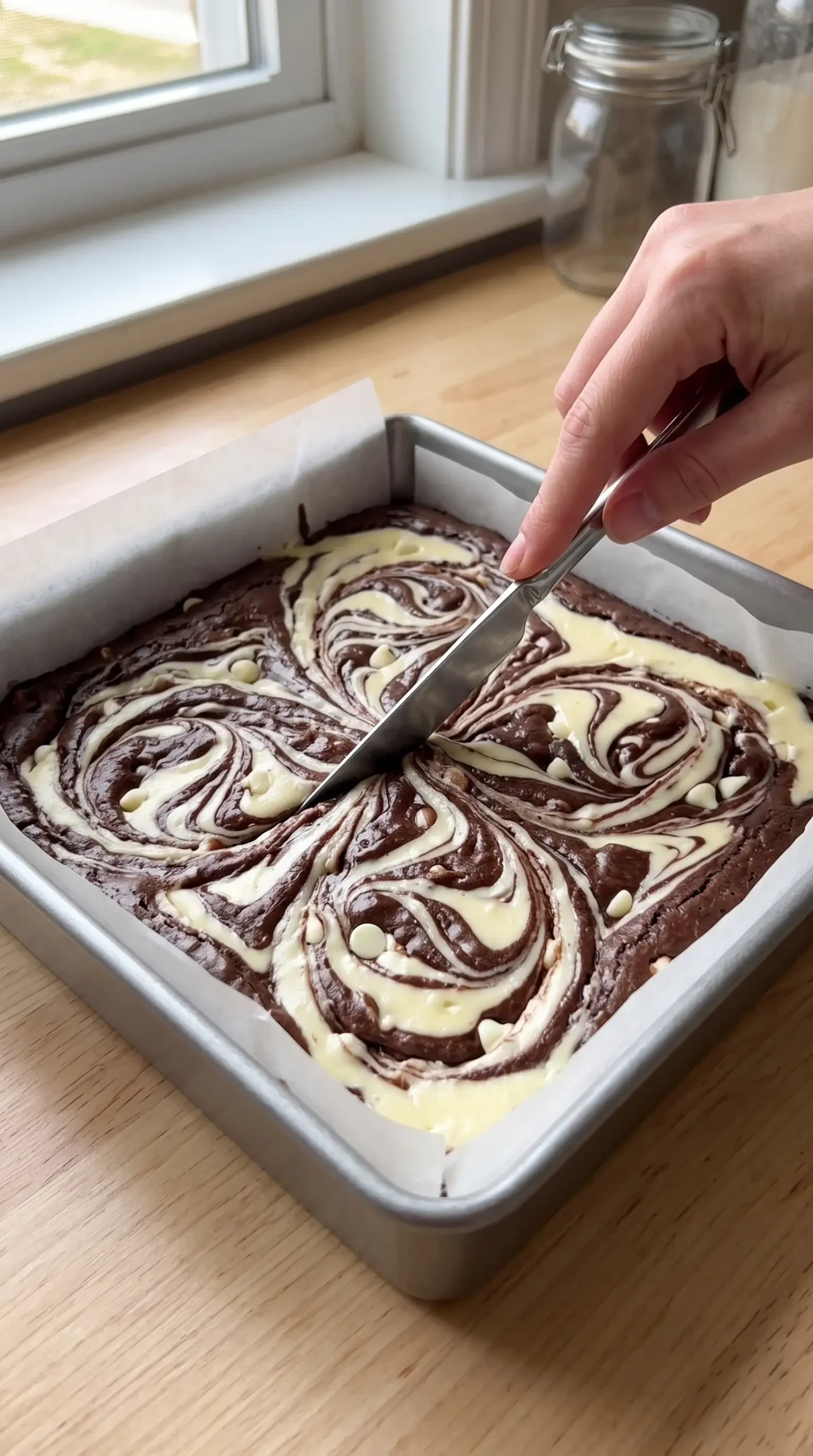 Knife swirling cheesecake topping through brownie batter in a baking pan